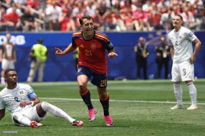 Spain's #21 celebrates a goal in red kit with pink boots, running on the field as an England player sits on the grass nearby.