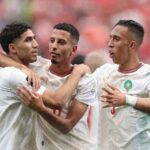 Three Morocco national team players in white jerseys with red and green trim celebrate together on the pitch The player on the left has his arm around the middle player while the player on the right gestures with his hands All three wear the Morocco crest and Puma logo A blurred stadium crowd in red fills the background  trybalnews