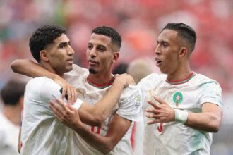 Three Morocco national team players in white jerseys with red and green trim celebrate together on the pitch. The player on the left has his arm around the middle player, while the player on the right gestures with his hands. All three wear the Morocco crest and Puma logo. A blurred stadium crowd in red fills the background.