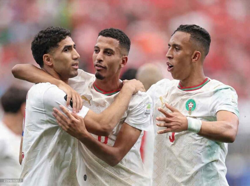 Three Morocco national team players in white jerseys with red and green trim celebrate together on the pitch The player on the left has his arm around the middle player while the player on the right gestures with his hands All three wear the Morocco crest and Puma logo A blurred stadium crowd in red fills the background  trybalnews