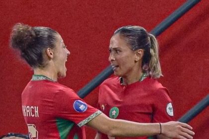 Two female soccer players in red-and-green uniforms hug and celebrate on the pitch.