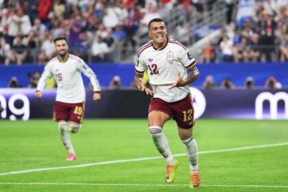 Spain defender Pedro Porro, wearing jersey number 12 in white and maroon, celebrates with arms outstretched and mouth open after scoring Spain's second goal in the FIFA World Cup 2026 Semi Final vs France at Dallas Stadium in Arlington, Texas. A teammate in jersey number 19 runs behind him on the pitch with a packed stadium in the background.