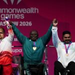 Nigerias Riluwan Idris centre wearing a green tracksuit and gold medal raises both arms and holds hands with the other medalists after winning gold in Mens Heavyweight Group A para powerlifting To his left Englands Matthew Harding in a white and red tracksuit with a silver medal holds a mascot To his right Indias Sudhir in a white tracksuit with a bronze medal also holds a mascot All three are on a podium at the 2026 Commonwealth Games in Glasgow  trybalnews