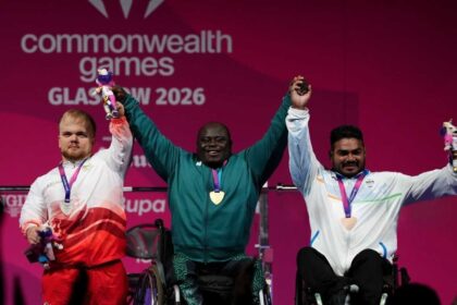 Nigeria's Riluwan Idris, centre, wearing a green tracksuit and gold medal, raises both arms and holds hands with the other medalists after winning gold in Men's Heavyweight Group A para powerlifting. To his left, England's Matthew Harding in a white and red tracksuit with a silver medal holds a mascot. To his right, India's Sudhir in a white tracksuit with a bronze medal also holds a mascot. All three are on a podium at the 2026 Commonwealth Games in Glasgow.