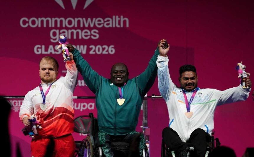 Nigerias Riluwan Idris centre wearing a green tracksuit and gold medal raises both arms and holds hands with the other medalists after winning gold in Mens Heavyweight Group A para powerlifting To his left Englands Matthew Harding in a white and red tracksuit with a silver medal holds a mascot To his right Indias Sudhir in a white tracksuit with a bronze medal also holds a mascot All three are on a podium at the 2026 Commonwealth Games in Glasgow  trybalnews