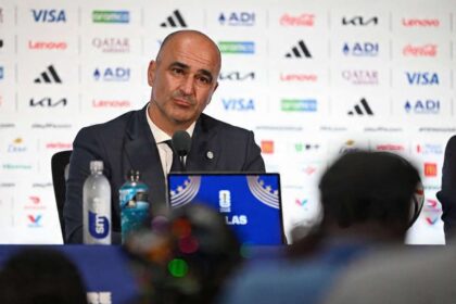 Roberto Martinez, head coach of Portugal, sits at a press conference table after the FIFA World Cup 2026 Round of 16 match against Spain at Dallas Stadium in Arlington, Texas on July 6, 2026. He wears a dark suit and tie in front of a sponsor backdrop with a microphone, water bottles, and a World Cup laptop in front of him.