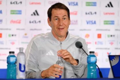 Male coach at a press conference, wearing a gray jacket with Argentina crest, speaking into a microphone with water bottles on the table.