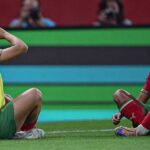 Two soccer players in red and green kits sit on the grass celebrating with fists to mouths during a match one wearing a yellow bib vest  trybalnews