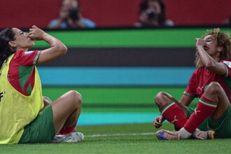 Two soccer players in red and green kits sit on the grass, celebrating with fists to mouths during a match, one wearing a yellow bib vest.