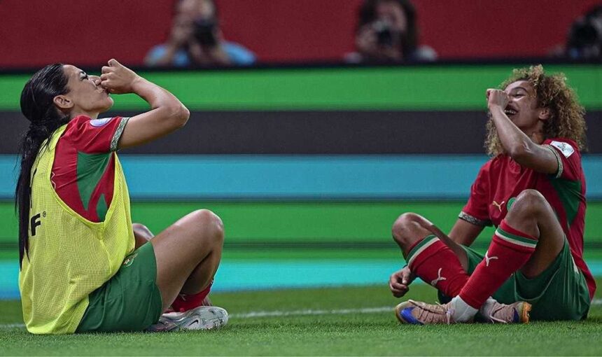 Two soccer players in red and green kits sit on the grass celebrating with fists to mouths during a match one wearing a yellow bib vest  trybalnews