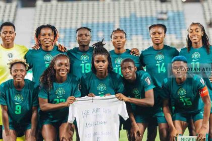 Nigerian women’s soccer team in teal uniforms posing for a group photo on the field, holding a white shirt with a get-well message.”] ,