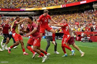 Ruben Vargas, #17 of Switzerland, celebrates with teammates after scoring the winning fifth penalty in a shootout during the FIFA World Cup 2026 Round of 16 match between Switzerland and Colombia at BC Place in Vancouver, British Columbia, Canada on July 7, 2026. Swiss players in red kits run and jump together on the pitch in front of a packed stadium.