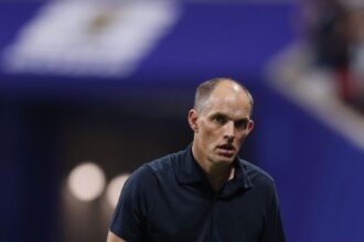 Man in a dark polo on a tennis court, looking focused against a blue background.