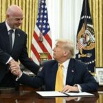 Two men in suits shake hands across a polished desk in the Oval Office with flags and a presidential seal in the background  trybalnews