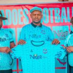 Three men in matching turquoise Niger Foods jerseys pose with a folded shirt at a sports event banner behind them promoting the team sponsor  trybalnews