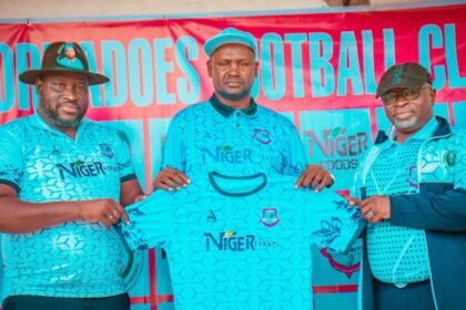 Three men in matching turquoise Niger Foods jerseys pose with a folded shirt at a sports event banner behind them, promoting the team sponsor.