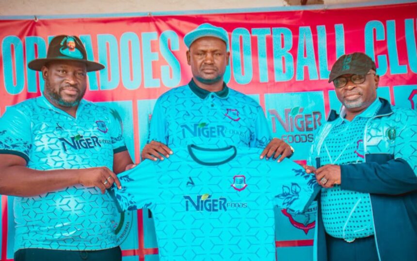 Three men in matching turquoise Niger Foods jerseys pose with a folded shirt at a sports event banner behind them promoting the team sponsor  trybalnews