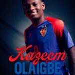 Portrait of a smiling man in a blue red football jersey with a crest featuring the name Kazem Olaigbe in red script and blue block letters in the foreground  trybalnews