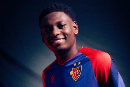 Portrait of a smiling man in a blue-red football jersey with a crest, featuring the name 'Kazem Olaigbe' in red script and blue block letters in the foreground.