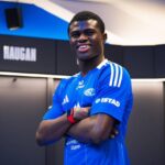 Young male athlete in a blue team jersey stands with arms crossed smiling in a locker room setting with name boards in the background  trybalnews