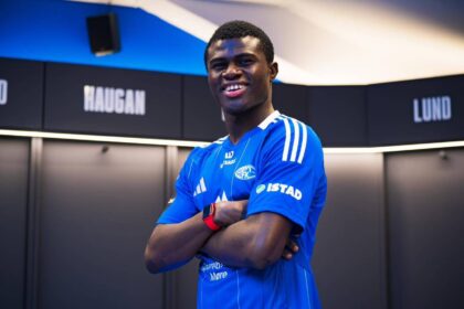 Young male athlete in a blue team jersey stands with arms crossed, smiling in a locker room setting with name boards in the background.
