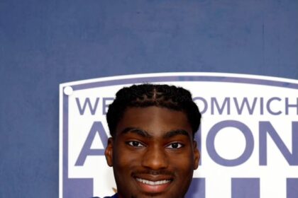 Smiling footballer in a blue West Bromwich Albion jersey signs a document at a table.
