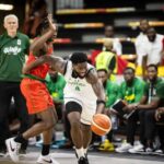 Nigerian player in a white jersey dribbles past a defender in red during a basketball game with teammates and spectators on the bench in the background  trybalnews
