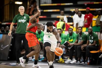 Nigerian player in a white jersey dribbles past a defender in red during a basketball game, with teammates and spectators on the bench in the background.