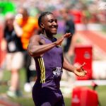 Male sprinter in a purple singlet finishes a track race smiling with arms raised in celebration as fans blur in the background  trybalnews