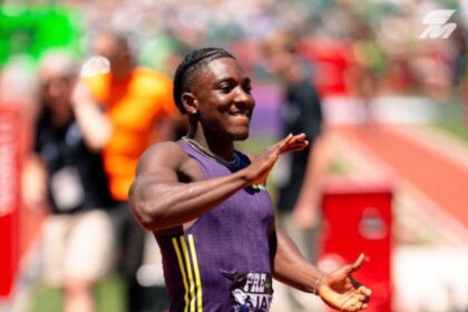 Male sprinter in a purple singlet finishes a track race, smiling with arms raised in celebration as fans blur in the background.