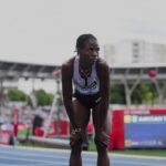 Female sprinter bending over at the track catching her breath during a race with stadium stands and scoreboard in the background  trybalnews
