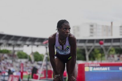 Female sprinter bending over at the track, catching her breath during a race with stadium stands and scoreboard in the background.