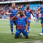Soccer players in blue and red striped uniforms celebrate on a grassy pitch the central player kneels and salutes while teammates stand behind with a cheering crowd in the background  trybalnews