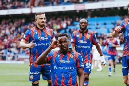 Soccer players in blue and red striped uniforms celebrate on a grassy pitch; the central player kneels and salutes while teammates stand behind, with a cheering crowd in the background.