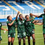 A soccer team in green uniforms celebrates together on the field raising arms in a victory cheer with blue stadium seats in the background  trybalnews