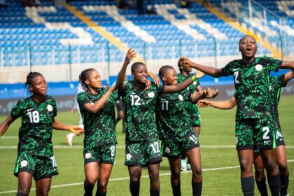 A soccer team in green uniforms celebrates together on the field, raising arms in a victory cheer with blue stadium seats in the background.