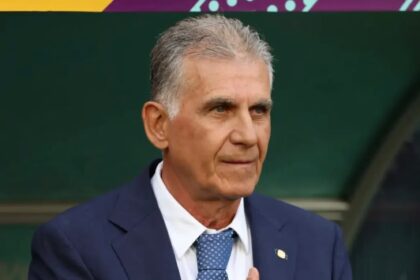 Older man in a navy suit and light-blue patterned tie, hands clasped, looking to the side against a green background.