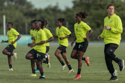 Group of female soccer players in neon training kits sprinting on a football field with a coach supervising.