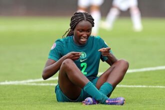 Female soccer player in a green jersey with number 2 sits on the grass, pointing to her chest.