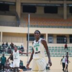Nigerian basketball player in a white jersey 9 stands on a gym court during a game with spectators and teammates in the background  trybalnews
