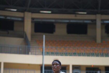 Nigerian basketball player in a white jersey #9 stands on a gym court during a game, with spectators and teammates in the background.