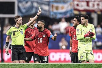 Soccer referee raises a yellow card as a red-clad player argues with teammates and a neon-clad goalie watches nearby on the field.