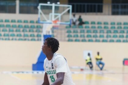Nigerian basketball player wearing #15 in white uniform, dribbling on an indoor court with a camouflage knee brace.