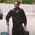Man in a black denim outfit pulls a wheeled suitcase while carrying a black duffel bag and a backpack outdoors  trybalnews