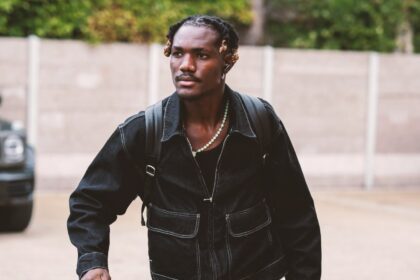 Man in a black denim outfit pulls a wheeled suitcase while carrying a black duffel bag and a backpack outdoors.