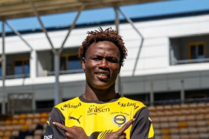 Soccer player in a yellow and black jersey crossing arms in a stadium, looking at camera with a smile.