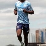 Soccer player in a light blue training jersey and navy shorts sprinting on a football pitch with a city skyline in the background  trybalnews