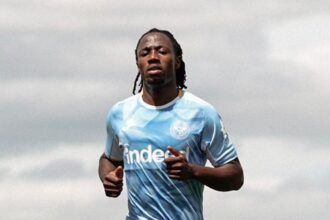 Soccer player in a light blue training jersey and navy shorts sprinting on a football pitch with a city skyline in the background