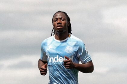 Soccer player in a light blue training jersey and navy shorts sprinting on a football pitch with a city skyline in the background