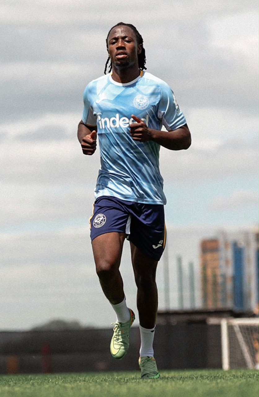 Soccer player in a light blue training jersey and navy shorts sprinting on a football pitch with a city skyline in the background  trybalnews